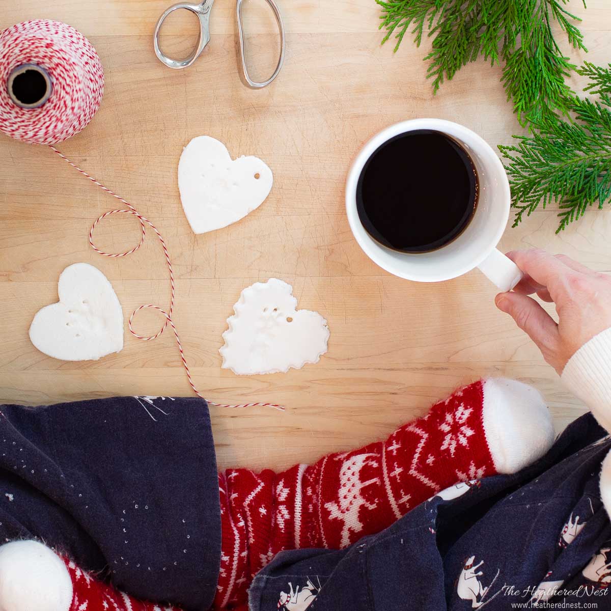 woman sitting cross-legged in Christmas pjs by a wood cutting board with three salt dough ornaments in heart-shapes. Also pictured butcher's twine, scissors and a cup of coffee