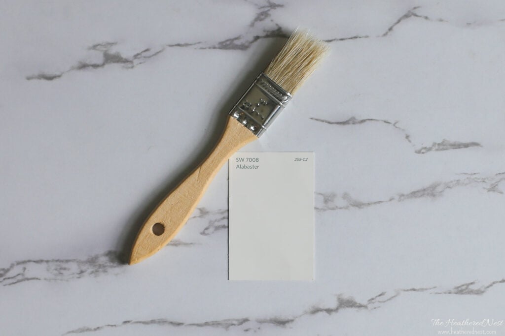 Sherwin Williams Alabaster SW 7008 paint chip on marble background with paintbrush