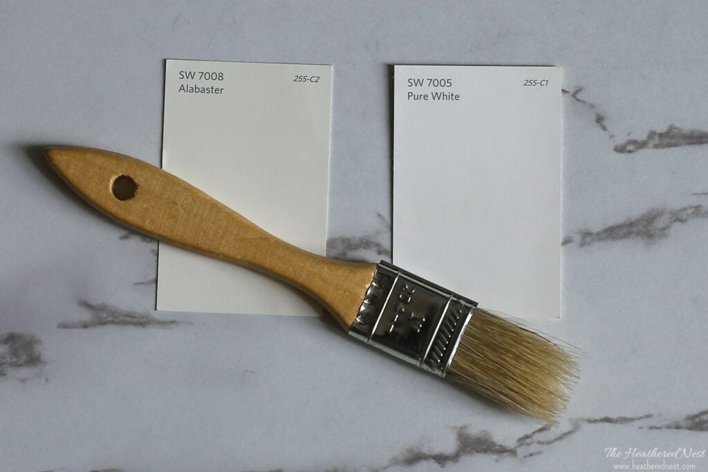 side by side paint swatch comparison of SW Alabaster 7008 and SW Pure White 7005 on a marble background with a wood paintbrush