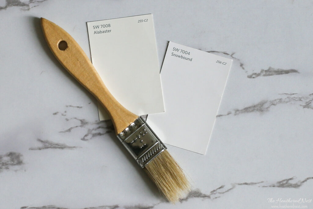 side by side paint swatch comparison of SW Alabaster 7008 and SW Snowbound 7004 on a marble background with a wood paintbrush